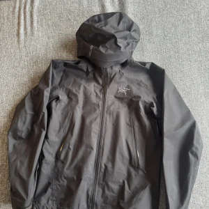 Svart vindjacka från Arc'teryx - Svart vindjacka från Arc'teryx med minimalistisk design och broderad logga på bröstet. Jackan har huva, hel dragkedja framtill och två snedställda fickor med dragkedja. Perfekt för dig som gillar stilren och funktionell outdoor-look.