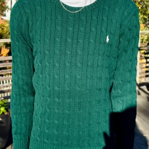 Grön kabelstickad tröja Polo Ralph Lauren - Mörkgrön kabelstickad tröja från Polo Ralph Lauren med klassisk rund halsringning och broderad vit logga på bröstet. Tröjan har lång ärm och ribbade muddar för en snygg passform. Perfekt för dig som gillar preppy och stilren look.