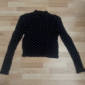 Svart prickig polotopp - Svart polotopp med vita prickar och långa ärmar. Toppen har en tight passform och är tillverkad i ett stretchigt material som sitter snyggt på kroppen. Perfekt för dig som gillar klassiska mönster med en modern twist. Storlek lapp avklippt.