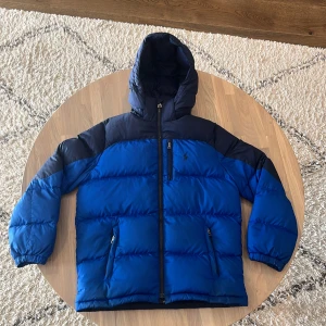 Blå pufferjacka från Polo Ralph Lauren - Säljer en blå och mörkblå pufferjacka från Polo Ralph Lauren med huva och dragkedja. Jackan har broderad logga på bröstet, två sidofickor med dragkedja och är riktigt varm och skön för kalla dagar. Perfekt för dig som vill ha en snygg och sportig vinterlook.