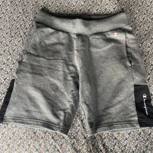 Grå Champion shorts med svart detalj - Snygga grå shorts från Champion med svart panel på sidorna och logotypen broderad i vitt. Mjuk och skön bomullsmix, perfekt för chill eller träning. Klassisk midja och två sidofickor. Enkel stil som funkar till det mesta.