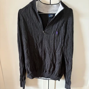 Mörkgrå kofta från Polo Ralph Lauren - Mörkgrå kofta från Polo Ralph Lauren med dragkedja framtill och diskret lila logga på bröstet. Tröjan är långärmad, har ribbade muddar och är tillverkad i mjukt material som passar perfekt till lager-på-lager-stil.