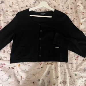 Svart kofta från Freequent, S - Svart kofta från Freequent i storlek S. Kortare modell med rund halsringning, knappar framtill och dekorativa fickor. Perfekt att slänga över en topp för en enkel och stilren look.