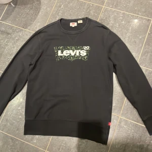 Svart Levi's sweatshirt  - Svart sweatshirt från Levi's med klassisk rund hals och lång ärm. På bröstet finns en stor Levi's-logga med grön leopardmönstrad bakgrund. Tröjan är i mjuk bomull och har en relaxed fit som passar perfekt till jeans eller joggers.