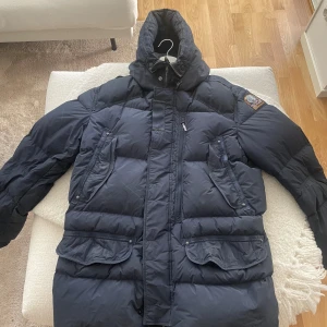 Mörkblå dunjacka Parajumpers XL - Säljer en mörkblå Parajumpers dunjacka i storlek XL. Jackan har huva, flera stora fickor och broderad patch på ärmen. Fylld med 90/10 dun/fjäder för max värme. Perfekt för kalla vinterdagar. Den passar XL-L-M, rekomenderar att jackan kemtvättas för att se ut som ny igen..