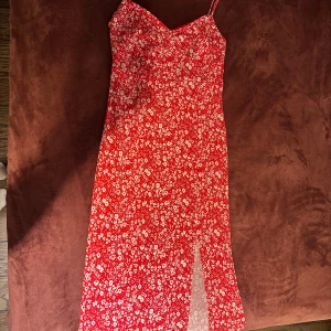 Röd blommig långklänning med slits - Superfin röd långklänning med vita blommor, smala axelband och en snygg slits framtill. Klänningen har figurnära passform. Materialet känns lätt och luftigt, och mönstret ger en riktigt söt vibe. Köparen står för frakten