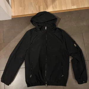 Moncler vindjacka - Svart vindjacka från Moncler med huva, hel dragkedja och två fickor med dragkedja framtill. Jackan har elastiska muddar och nederkant samt Moncler-logga på ärmen. Lätt och smidig jacka i syntetmaterial, perfekt för blåsiga dagar. Ny skick och tagen finns kvar