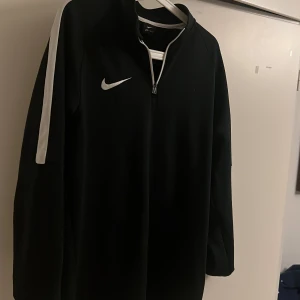 Svart Nike Dri-Fit half zip tröja - Svart långärmad Nike Dri-Fit tröja med vit dragkedja och vita detaljer längs ärmarna. Perfekt för träning eller chill dagar. Swoosh-logga på bröstet och sportig passform. Materialet är lätt och andas, vilket gör den skön att ha på sig.