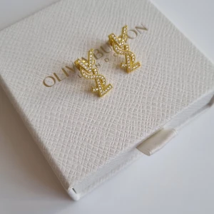 YSL örhängen i guld med strass - Snygga örhängen i guldton med glittrande strassdetaljer. Ikonisk logga i stilren design, perfekta för att lyfta din look. Stiftmodell som sitter bekvämt på örat och ger en lyxig känsla. Lådan ingår ej.
