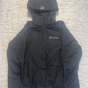 Svart vindjacka från Tenson - Svart vindjacka från Tenson med huva, dragkedja och praktiska fickor. Jackan är tillverkad i polyester och har meshfoder på insidan. Perfekt för blåsiga dagar och enkel att matcha med allt.