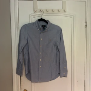 Blå skjorta från Ralph Lauren - Klassisk blå skjorta från Ralph Lauren med broderad logga på bröstet. Skjortan har lång ärm, knappar framtill och button down-krage. Tillverkad i 100% bomull för en skön och stilren känsla.