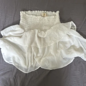 Vit volangkjol från Chelsea XS - Supersöt vit kortkjol från Chelsea med smockad midja och volangdetaljer. Kjolen har en luftig och flowy känsla, perfekt för varma dagar. Den elastiska midjan gör den bekväm och lätt att bära. Styla med en crop top eller en enkel t-shirt för en fräsch look.