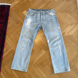 Levi's 501 ljusblå raka jeans - Ljusblå Levi's 501 jeans med klassisk rak passform och femficksdesign. Jeansen har en vintage vibe med slitningar och vita färgstänk på flera ställen. Tillverkade i 100% bomull för en autentisk jeanskänsla. Perfekta för en avslappnad och trendig look. Även uppsydda så den sanna storleken skulle jag säga är 32/32