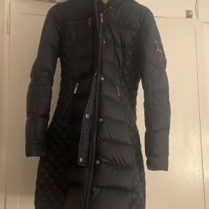 Mörkblå lång dunjacka med huva - Lång mörkblå dunjacka från rock and blue, pris kan diskuteras. Storlek xs men passar även s , jackan har ett hål i fickan men inget som syns.det är bara att sy ihop de