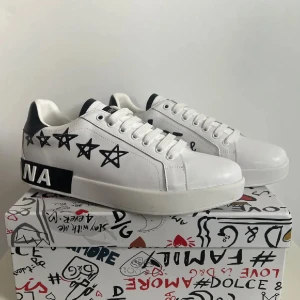 Dolce & Gabbana vita sneakers med stjärnor - Snygga vita sneakers från Dolce & Gabbana med svarta stjärnor på sidorna och svart detalj vid hälen. Skorna har snörning, platt sula och coola grafiska detaljer på sulan och lådan. Perfekta för dig som vill sticka ut med en unik och trendig look.