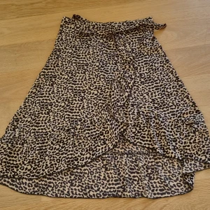 Leopardmönstrad omlottkjol Neo Noir - Snygg omlottkjol från Neo Noir med leopardmönster i beige och svart. Kjolen har volangdetaljer längs nederkanten och knytning i midjan. Perfekt för dig som vill sticka ut med en trendig och cool look.