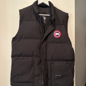 Svart dunväst från Canada Goose - Svart dunväst från Canada Goose med klassisk logga på bröstet och två stora fickor framtill. Inköpt från Care Of Carl. Använd 2 höst säsonger. Kvitto finns för autenticitet. 