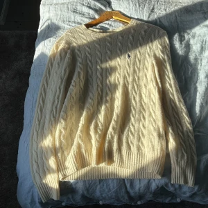 Kabelstickad vit tröja Polo Ralph Lauren - Kabelstickad vit tröja från Polo Ralph Lauren med klassisk rund hals och broderad marinblå logga på bröstet. Tröjan är i mjuk ullblandning och har ribbade muddar vid ärmslut och nederkant. Perfekt för lager-på-lager under kyliga dagar.