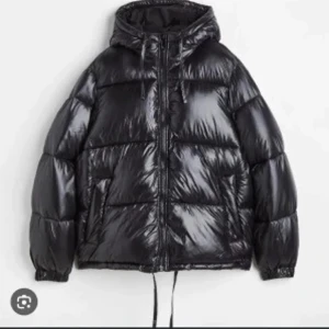 Svart pufferjacka med huva - Säljer en svart, glansig pufferjacka med huva och dragkedja framtill. Jackan har en oversized passform, snörning nertill och långa ärmar. Perfekt för kalla dagar när du vill vara både varm och snygg.