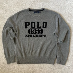 Grå tröja Polo Ralph Lauren | M - Grå tröja från Polo Ralph Lauren med svart broderad text 'POLO 1967 ATHL. DEPT.' på bröstet. Klassisk rund halsringning, ribbade muddar och nederkant. Skön och stilren, perfekt till jeans eller chinos.