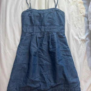 Blå jeansklänning med smala axelband - Snygg blå jeansklänning med smala axelband och markerad midja. Klänningen har en kort längd och en synlig dragkedja baktill. Materialet är klassisk denim och modellen är ärmlös med lätt utställd kjol. Perfekt för dig som gillar en enkel men cool stil.