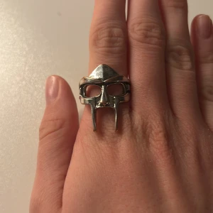 Silverfärgad ring med maskdesign - Unik ring i silverfärgad metall formad som en mask, inspirerad av klassiska hjälmar. Ringen har detaljerade utskärningar för ögon och näsa, vilket ger en cool och edgy vibe. Perfekt accessoar för dig som vill sticka ut och addera karaktär till din stil. STOR I STORLEKEN 