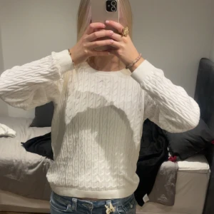 Vit kabelstickad tröja - Säljer en vit kabelstickad tröja med rund hals och långa ärmar. Tröjan har klassiskt mönster och ribbade muddar vid ärmslut och nederkant. Perfekt att styla med jeans för en clean look.