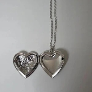 Halsband med hjärtformat medaljong 45 cm - Snyggt silverfärgat halsband med hjärtformat medaljong som går att öppna. Medaljongen har graverade detaljer på framsidan och plats för en liten bild eller minne inuti. Kedjan är tunn och justerbar för perfekt passform.