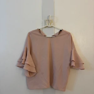 Ljusrosa blus med volangärmar - Säljer en ljusrosa blus med snygga volangärmar och rund halsringning. Blusen har en lös passform och är perfekt för dig som gillar en stilren look med en twist. Materialet känns mjukt och lätt, vilket gör den skön att bära. Passar till både jeans och kjol.