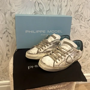 Philippe Model vita sneakers i skinn - Snygga vita Philippe Model sneakers i skinn med grå detaljer och snörning. Skorna har en klassisk låg siluett, rund tå och kontrasterande blå insida. Perfekta för dig som gillar en trendig och avslappnad look. Har sina skador se bild. 