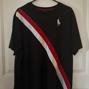 Svart t-shirt Polo Ralph Lauren Youth XL, 170 - Svart t-shirt från Polo Ralph Lauren i storlek Youth XL, strlk 170 (18-20) med diagonal röd och vit rand över framsidan och klassisk tryckt logga på bröstet. 100% polyester, Ralph Laurence Performance. Ej använd. 