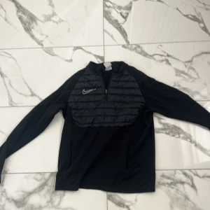 Svart Nike Therma-Fit half zip tröja - Nike Therma-Fit tröja i svart med quiltad frontpanel och halv dragkedja. Swoosh-logga på bröstet och gråa detaljer längs ärmarna. Perfekt för träning eller chill dagar. Långärmad och tillverkad i funktionsmaterial som håller dig varm Använd under 3 tillfällen , var för liten.