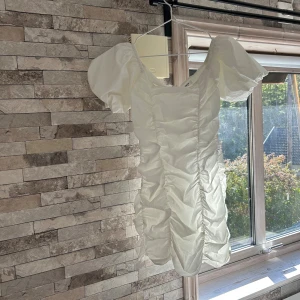 Vit puffärm klänning från Glamorous - Supersöt vit kort klänning från Glamorous med puffärmar och snygg rynkad effekt framtill. Klänningen har dragkedja i ryggen och är tillverkad i ett lätt, glansigt material som känns lyxigt. Perfekt för dig som vill sticka ut med en feminin och trendig look.