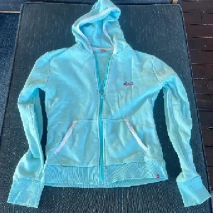 Ljusblå hoodie från Esprit - Ljusblå hoodie från Esprit med dragkedja framtill och broderad logga på bröstet. Tröjan har huva, långa ärmar och ribbade muddar samt fickor med vita detaljer. Tillverkad i mjuk bomullsmix, perfekt för chill dagar.