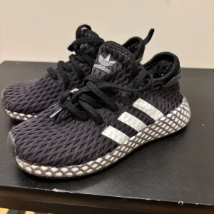 Svarta Adidas sneakers med nätmönster - Säljer ett par svarta Adidas sneakers med vit sula och klassiska tre ränder på sidan. Ovandelen har ett coolt nätmönster och skorna har snörning. Perfekta för dig som gillar streetstyle och vill ha något unikt på fötterna.