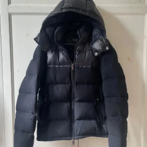 Svart pufferjacka Polo Ralph Lauren - Snygg svart pufferjacka från Polo Ralph Lauren med huva och dragkedja. Jackan har en mix av glansigt och matt material, quiltad design och två fickor med dragkedja. Perfekt för kalla dagar när du vill hålla stilen. Jackan Säljs ej längre därav priset 