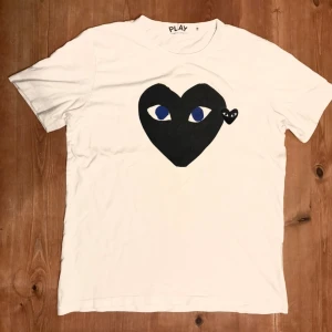 Vit Comme des Garçons Play t-shirt - Vit t-shirt från Comme des Garçons Play med det ikoniska svarta hjärta-trycket med ögon på bröstet. Klassisk passform och rund hals. Perfekt för dig som gillar grisch stilen och vill sticka ut med en cool detalj. PASSAR STORLEK S
