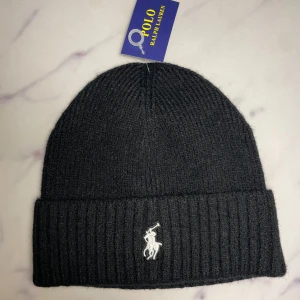 Svart stickad mössa Polo Ralph Lauren - Svart ribbstickad mössa från Polo Ralph Lauren med klassisk broderad logga framtill. Mjuk och varm, perfekt för kyliga dagar. Enkel och stilren design som passar till allt. Tveka inte att höra av er vid frågor/prisförslag!🙌