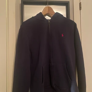 Mörkblå hoodie från Ralph Lauren - Säljer en mörkblå hoodie från Ralph Lauren med dragkedja och klassisk röd broderad logga på bröstet. Tröjan har huva, fickor framtill och är tillverkad i mjuk bomull. Perfekt för en chill och stilren look.
