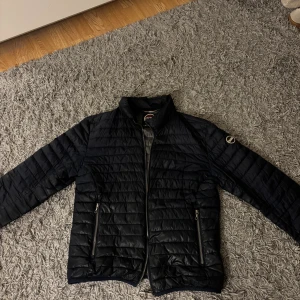 Svart pufferjacka från Colmar - Snygg svart pufferjacka från Colmar med quiltad design och raka linjer. Jackan har dragkedja framtill, två dragkedjefickor och klassisk Colmar-logga på ärmen. Perfekt för kyliga dagar och enkel att matcha med allt.