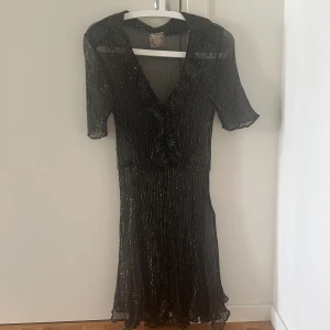 Svart glittrig klänning med v-ringning - Svart klänning från line of oslo med glittriga ränder och v-ringad hals. Klänningen har korta ärmar och är tillverkad i ett lätt, transparent material med ett snyggt fall. Perfekt för dig som vill sticka ut med lite extra glitter. Passar som snygg cover up på strandklubb. Står xl men skulle snarare säga S