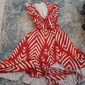 Röd och vit omlottklänning med mönster - Säljer en snygg omlottklänning med röd och vit randigt mönster. Klänningen har v-ringad hals, volangdetaljer vid axlarna och medföljande bälte i samma tyg. Perfekt för dig som gillar färgstarka och unika plagg.