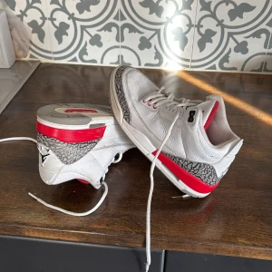 Nike Air Jordan 3 Retro White/Red - Nike Air Jordan 3 Retro sneakers i vitt skinn med röda och grå detaljer samt ikoniskt elephant print runt hälen. Skorna har klassisk snörning. Ett snöre är lite längre en det andra. Insidan är fodrad i rött tyg och yttersulan har tydlig Jordan-logga. Perfekt för dig som gillar streetwear och sneakerskultur.