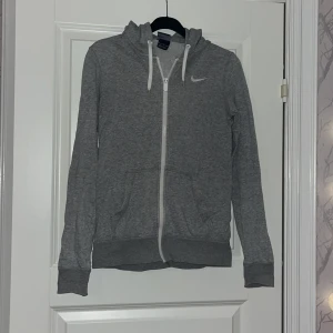 Grå hoodie från Nike med dragkedja - Grå hoodie från Nike med vit dragkedja och snören i huvan. Klassisk Nike-logga på bröstet, två fickor framtill och ribbade muddar. 
