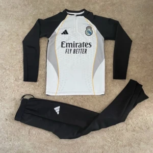 Real Madrid långärmad träningströja Adidas - Säljer en långärmad Real Madrid träningströja från Adidas i svart och vitt med gula detaljer. Tröjan har klubbmärke och sponsortryck framtill samt dragkedja vid halsen. Materialet är lätt och andas, perfekt för fotbollsträning.
