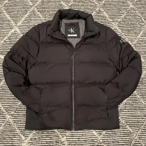 Svart pufferjacka från Calvin Klein Jeans - Säljer en svart pufferjacka från Calvin Klein Jeans med stor logga på ärmen. Jackan är vadderad med dun och fjäder, har dragkedja framtill och två fickor med dragkedja. Perfekt för kalla dagar. Riktigt bra skick, i procip nyskick.
