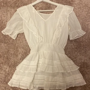 Vit volangklänning med kort ärm - Superfin vit kortklänning med volanger och smock i midjan. 💞 Klänningen har korta ärmar med volangkant, v-ringning fram och flera lager volanger i kjolen! Tillverkad i ett lätt och luftigt tyg, perfekt för varma dagar. Säljer då den är för liten, har bara används en gång, superbra skick. 🤍 Klänning är ej tillgänglig i butik längre. ❤️