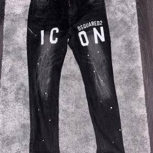 Svarta Dsquared2 ICON jeans, stl 48 - Snygga svarta jeans från Dsquared2 med stor vit 'ICON'-print framtill och coola vita färgstänk över hela byxorna. Jeansen har raka ben, klassiska fickor och en unik streetkänsla. Perfekta för dig som vill sticka ut med en trendig look.