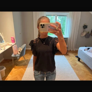 Svart blus med spetsärmar - Supersnygg svart blus med volangiga spetsärmar och rund halsringning. Toppen har ribbad struktur i tyget och detaljer i spets både vid axlar och hals. Perfekt att styla med jeans eller kjol för en trendig look.