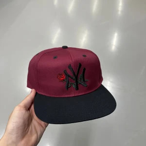 Bordeaux New York Yankees keps - Snygg bordeaux keps från New York Yankees med svart skärm och broderad logga framtill, dekorerad med en röd ros. Justerbar snapback baktill och grön undersida på skärmen. Perfekt accessoar för att lyfta din streetwear-look.